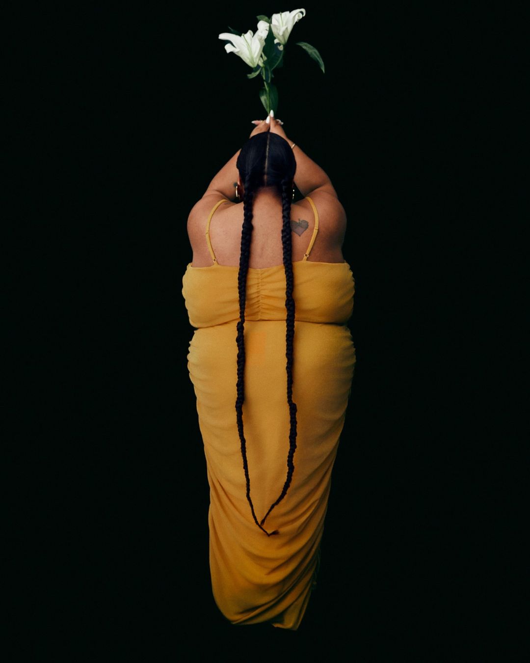 Woman in a yellow dress holding white lilies against a black background. The shot is from above, showing braids and dress details.