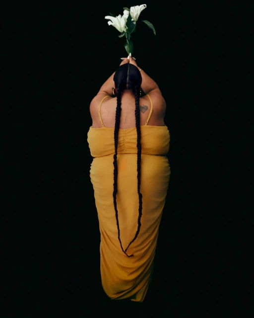 Woman in a yellow dress holding white lilies against a black background. The shot is from above, showing braids and dress details.