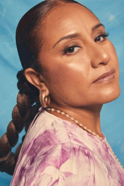 Close-up portrait of an adult woman with a braided ponytail, gold hoop earrings, and a patterned top against a blue background.