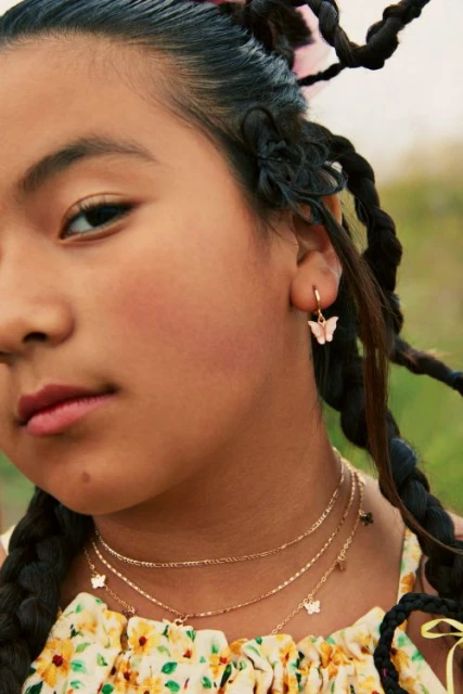 Close-up of a girl with braided hair wearing butterfly earrings and a layered necklace with butterfly charms outdoors.
