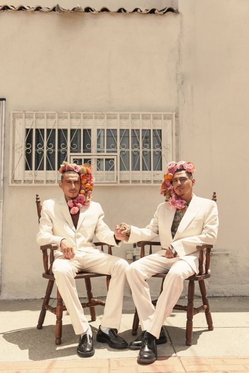 Two men in white suits and flower headpieces sit side-by-side on a wooden chair outdoors against a white wall with a barred window.
