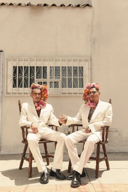 Two men in white suits and flower headpieces sit side-by-side on a wooden chair outdoors against a white wall with a barred window.