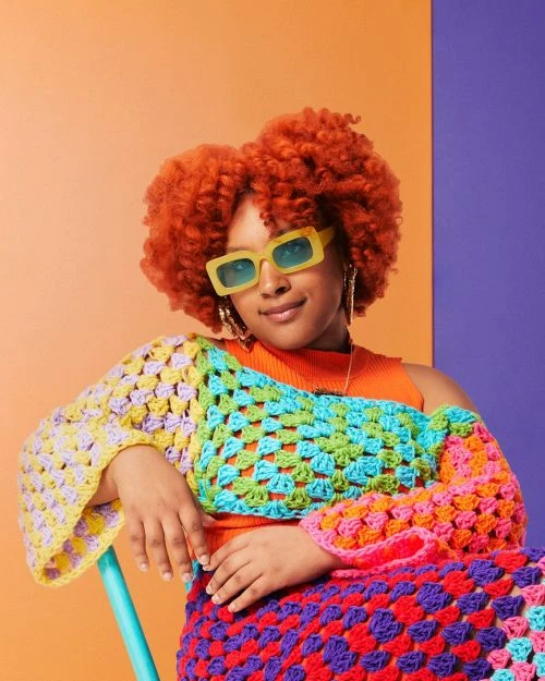Portrait of a smiling woman with curly red hair wearing retro yellow sunglasses and a colorful crochet top against a vibrant background.