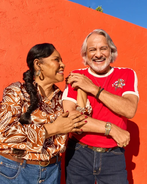 Smiling couple poses against an orange wall, woman in patterned top and man in a red shirt with jeans, capturing a cheerful outdoor moment.