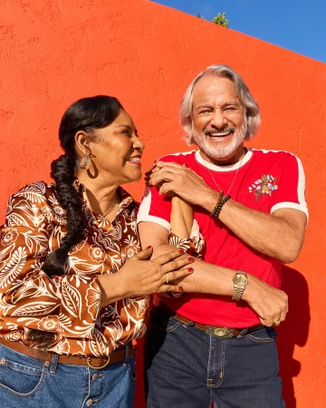Smiling couple poses against an orange wall, woman in patterned top and man in a red shirt with jeans, capturing a cheerful outdoor moment.