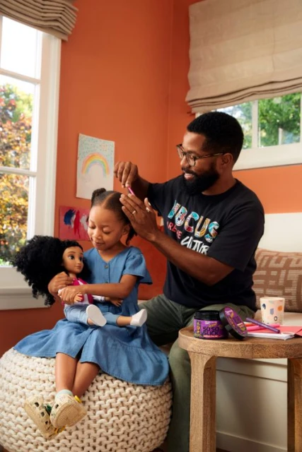 Father styling his daughter's hair as she sits with a doll. They're sitting on a white pouf, with a stool nearby in a brightly lit room.