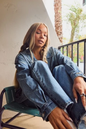 Woman with braided blonde hair wearing a denim jacket and jeans seated on a teal chair on a balcony.