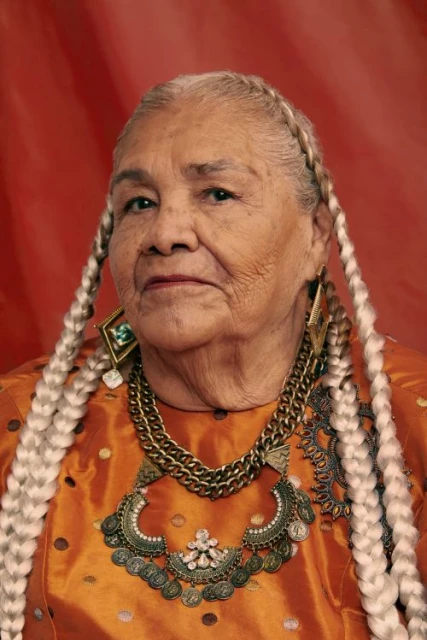 Portrait of an elderly woman with fair skin and long braided hair, wearing a gold and bronze colored silk top and ornate metal jewelry.