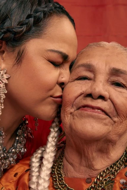 Elderly Hispanic woman with bright skin and white braided hair is embraced by a younger woman wearing gemstone jewelry.