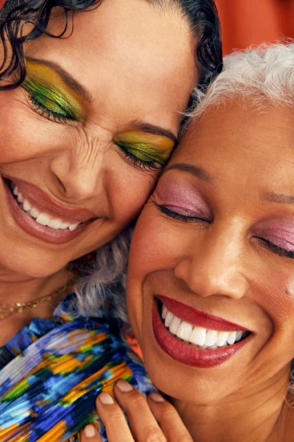 Close-up of two smiling women with eyes closed; one with colorful eyeshadow and one with red lipstick share an affectionate moment.
