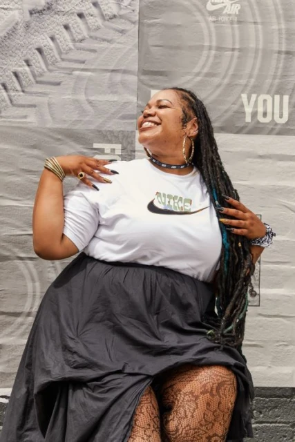 Woman with braids smiles against a textured grey backdrop wearing a white Nike t-shirt, gold jewelry, and grey skirt with lace tights.
