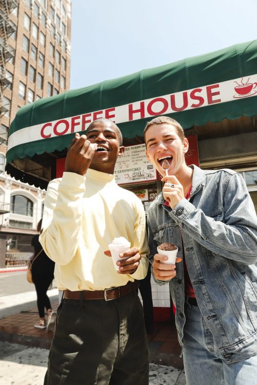 Two young adults stand outside a coffee shop, each enjoying a frozen treat on a sunny day with a city building in the background.