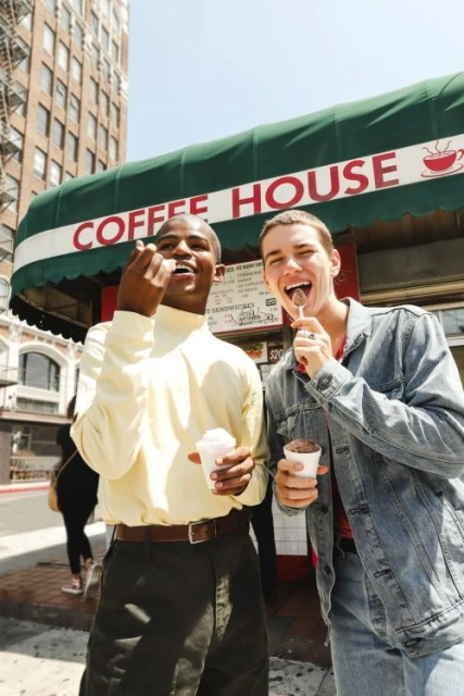 Two young adults stand outside a coffee shop, each enjoying a frozen treat on a sunny day with a city building in the background.