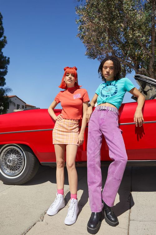 Two young adult models pose in front of a classic red car on a sunny day, wearing colorful vintage-inspired clothing and accessories.