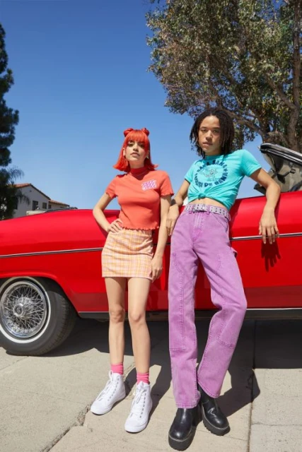 Two young adult models pose in front of a classic red car on a sunny day, wearing colorful vintage-inspired clothing and accessories.