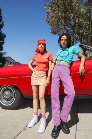 Two young adult models pose in front of a classic red car on a sunny day, wearing colorful vintage-inspired clothing and accessories.