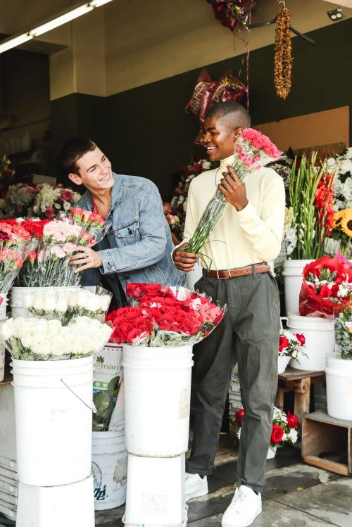 Two men at a flower shop, one holding a bouquet and the other arranging flowers in buckets; a cheerful, candid lifestyle moment.