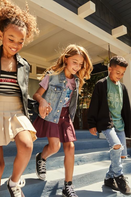 Three smiling children, two girls and a boy, walk down the stairs outdoors wearing casual outfits and sneakers in a medium shot.