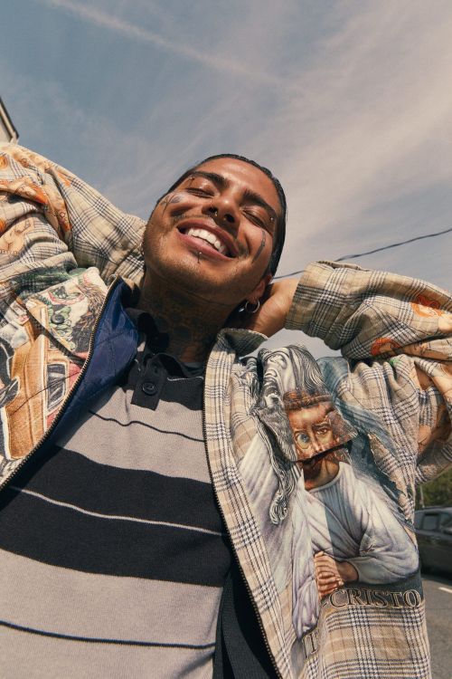 Smiling adult male wearing a patterned jacket with artwork, posing outdoors under a bright sky, showcasing fashion style and design.