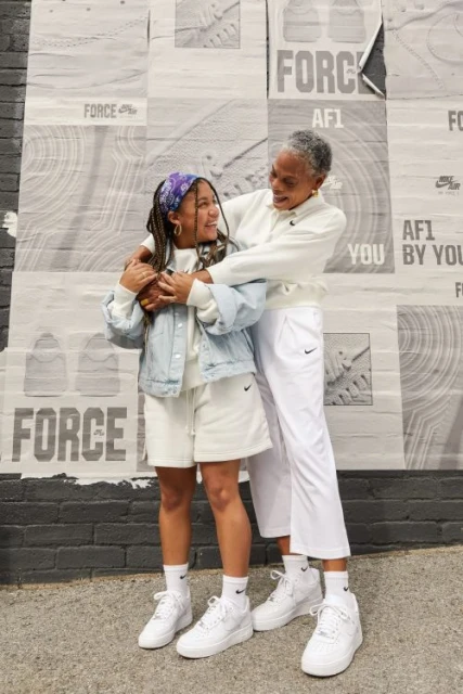 Two generations of women embrace in front of a wall covered in vintage Nike Force posters, both wearing Nike shoes and clothing.