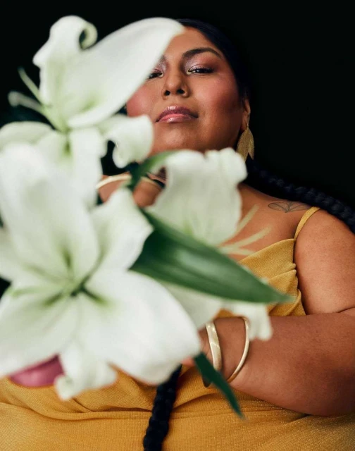 Portrait of an adult woman with braided hair in a yellow dress, holding a white lily against a dark background in a beauty shot.