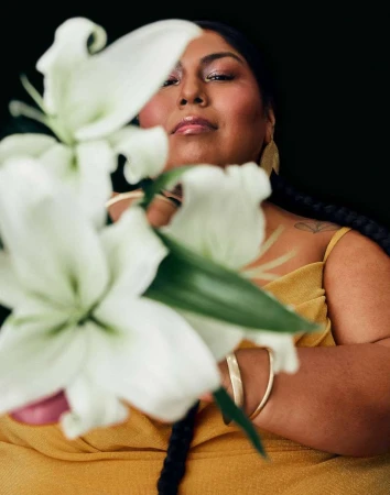 Portrait of an adult woman with braided hair in a yellow dress, holding a white lily against a dark background in a beauty shot.