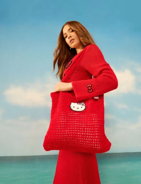 Woman in red knitwear poses with a red open-weave Hello Kitty tote bag against a backdrop of blue sky and ocean.