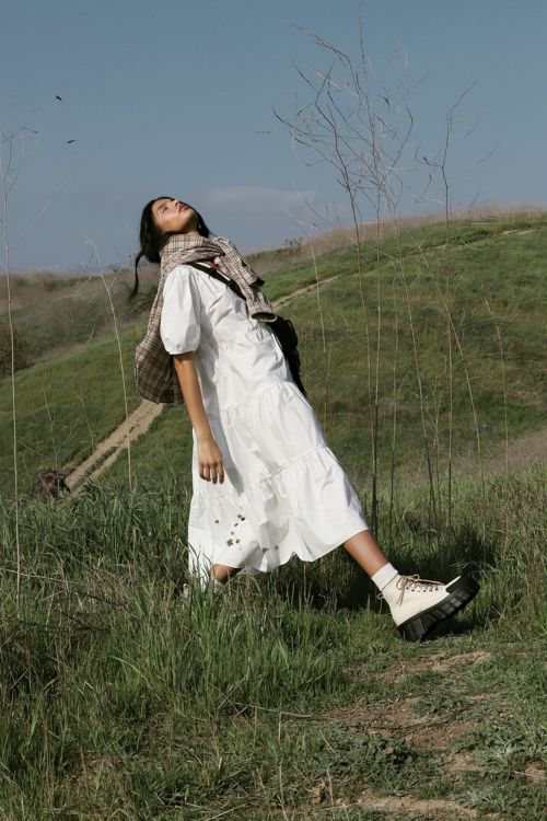 Woman in a white midi dress and platform boots standing outdoors on a grassy hill under a clear sky.