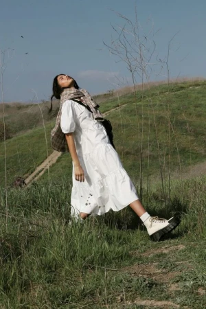 Woman in a white midi dress and platform boots standing outdoors on a grassy hill under a clear sky.