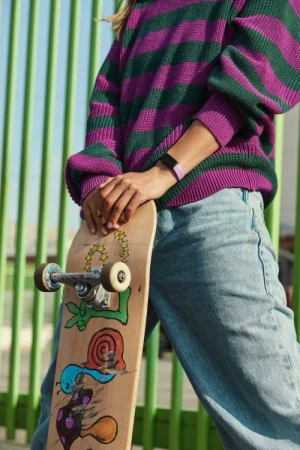 A female in a striped sweater leans on a skateboard with colorful drawings, set against a green fence in an outdoor setting.