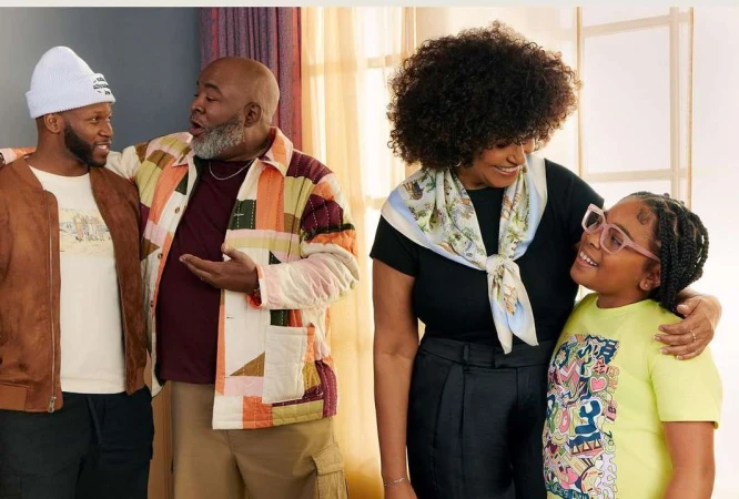 Indoor portrait of a multi-generational family of five, featuring two adult men, an adult woman, and a girl and a young child, smiling.
