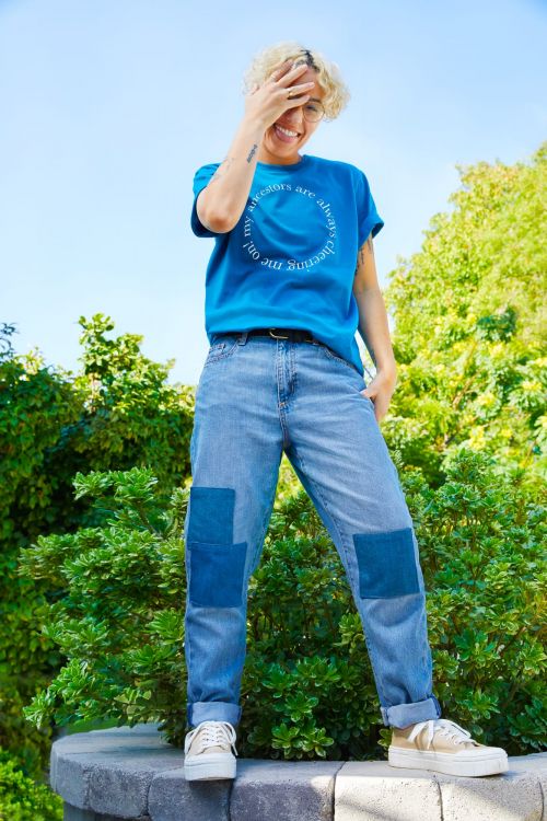 An adult woman with blonde hair stands smiling in blue jeans and a tee shirt with white text, outdoors with green trees in the background.