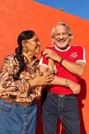 An older couple laughing and holding arms in front of a bright orange textured wall outdoors.