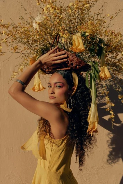Woman in yellow dress carrying a basket of yellow flowers on her head, standing against a tan wall in soft, diffused lighting.