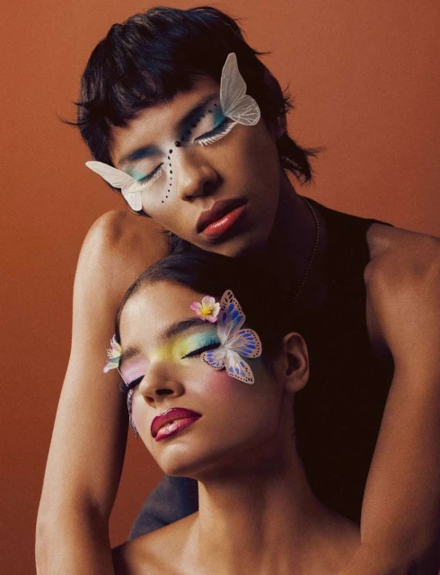 Two people with decorative eye makeup including butterflies, against a bronze background in a beauty style fashion portrait.