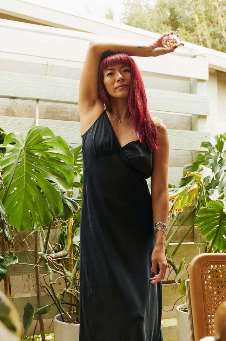 Woman in black dress poses in outdoor setting with monstera plant, filtered light, and a natural, relaxed aesthetic.