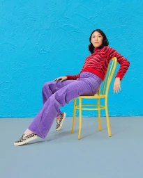 Woman in a red patterned sweater and purple corduroy pants seated on a yellow chair against a blue textured backdrop.