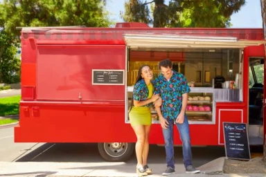 Man and woman standing next to a bright red food truck parked on a sunny Los Angeles street.