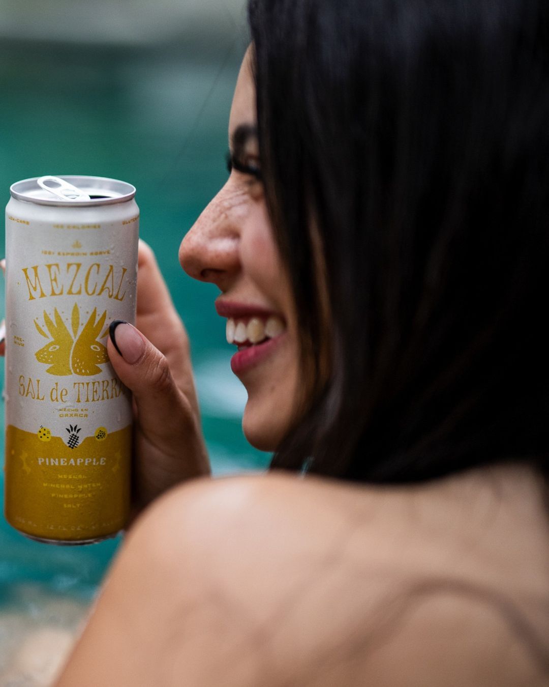 Smiling woman holds a can of Mezcal Sal de Tierra Pineapple drink against a pool backdrop, showcasing a relaxed and refreshing scene.
