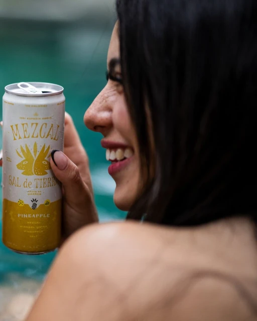 Smiling woman holds a can of Mezcal Sal de Tierra Pineapple drink against a pool backdrop, showcasing a relaxed and refreshing scene.