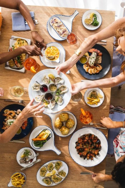 Overhead view of a group of people dining at a light wood table laden with seafood dishes and drinks, creating a vibrant culinary scene.
