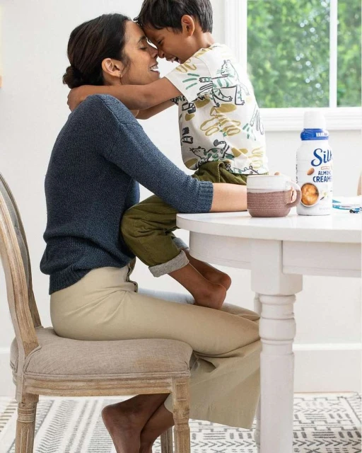 A mother and child embrace warmly indoors near a window, with a carton of Silk Almond Creamer and a mug on a white table.