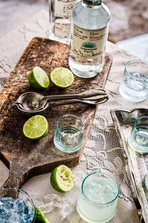 Still life shot of Casamigos tequila with limes and cocktail glasses on a wooden board and patterned tablecloth, suggesting a cocktail setting.