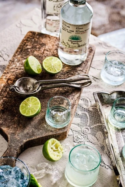 Still life shot of Casamigos tequila with limes and cocktail glasses on a wooden board and patterned tablecloth, suggesting a cocktail setting.