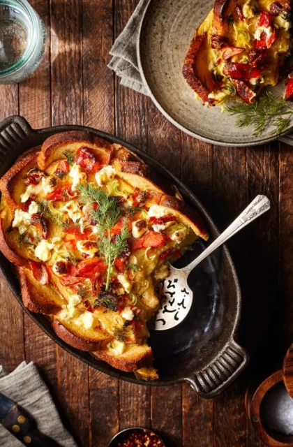 Overhead shot of a baked breakfast casserole featuring bread, eggs, cheese, and vegetables, served in a cast iron dish on a wood table.