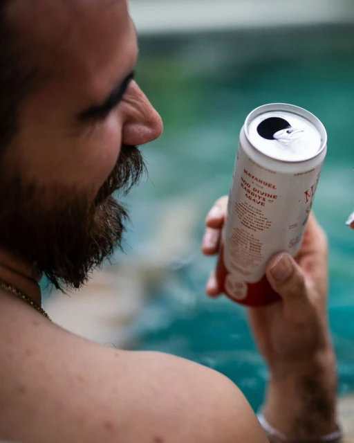 Man with beard holds a can of beverage outside. He is by a body of water. The can has a white background and red lettering.