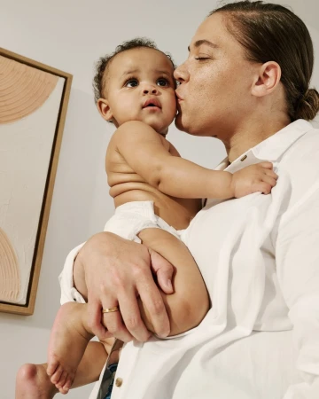 A woman in a white shirt kisses a baby on the cheek indoors, with an art piece visible in the background and neutral color palette.