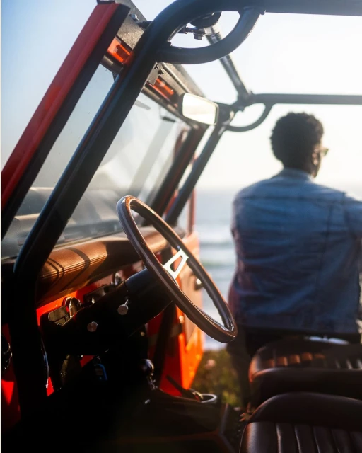 An adult male stands near an orange Jeep parked on a grassy hill overlooking the ocean on a bright day, captured from behind the vehicle.