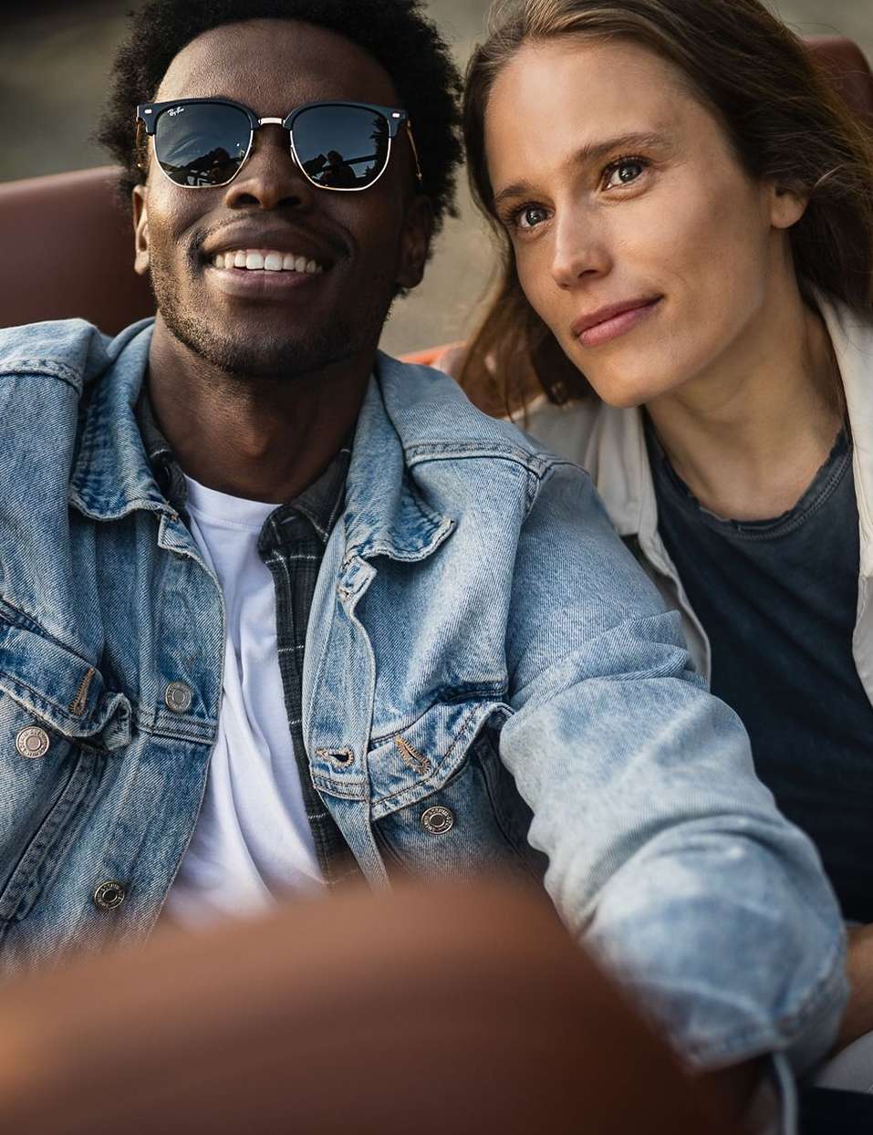 Close-up of a smiling African-American man wearing sunglasses, sitting next to a Caucasian woman in casual outfits.