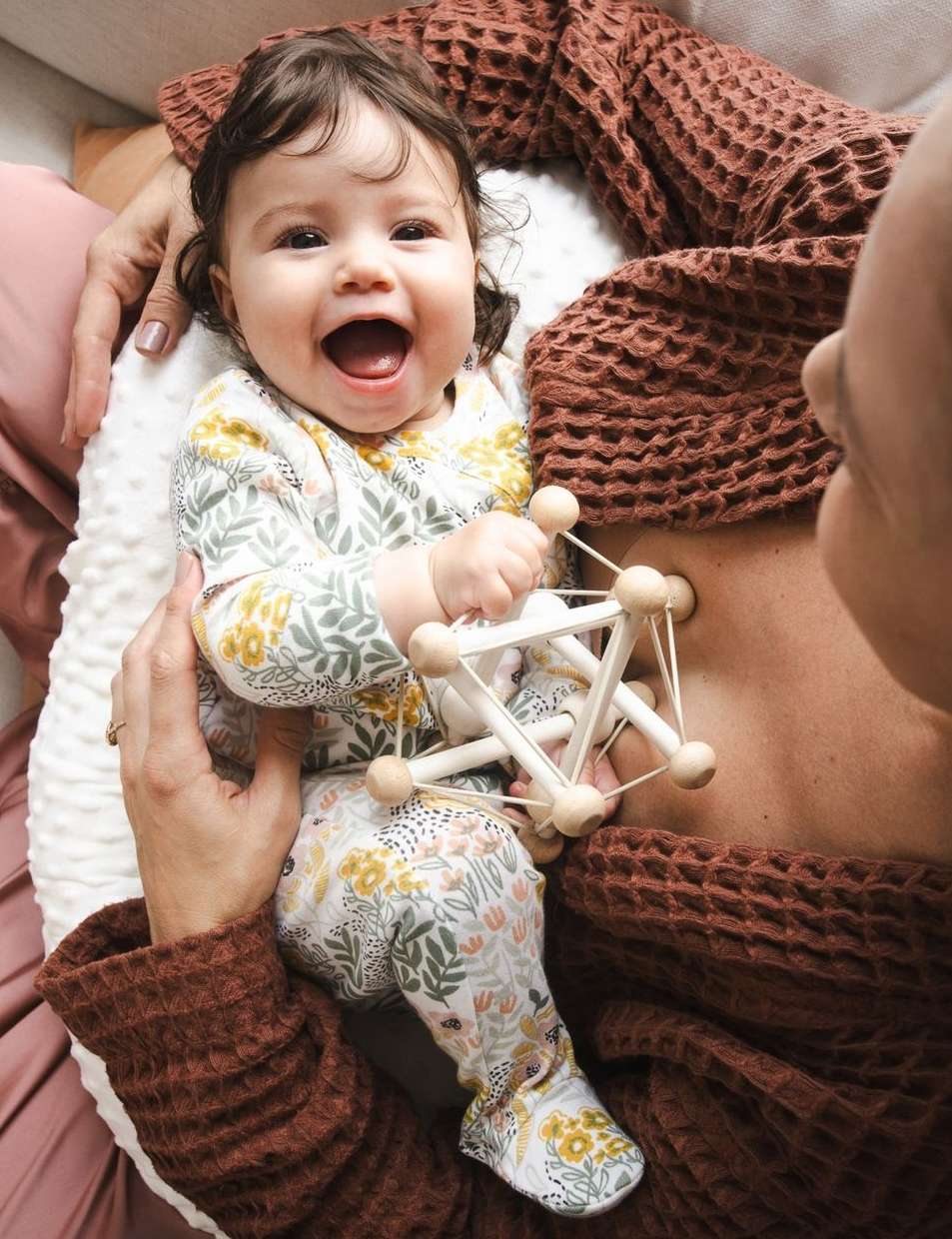 Happy baby in floral print outfit lies next to a woman wearing a brown waffle-knit top while holding a wooden geometric toy.
